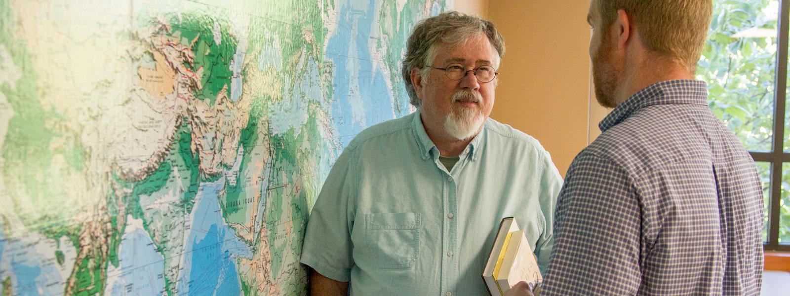 ministry professor holds books and talks to student while standing in front of world map on wall